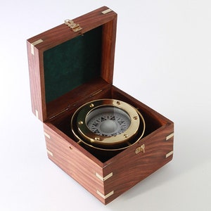 May include: A brass compass with a white face and black markings, in a wooden box with a hinged lid. The box is made of dark wood with brass accents.