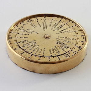 May include: A gold-toned metal world time zone clock with a rotating dial. The dial features city names and time zones around the perimeter. The clock is a useful tool for travellers and those who work with international time zones.