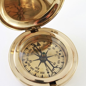 May include: A brass compass with a white face and black markings. The compass is open and the needle is pointing north. The compass is 80_100 120 140 E SE 180 NE 20 40 60 SW W 200 220 240 260 280 300 320 S NW.