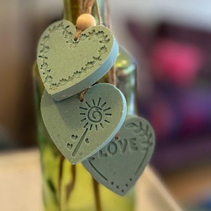 May include: Three sage green heart-shaped ornaments made of a matte material, hanging from twine with a small wooden bead. One heart features a floral border, one a sun-like graphic, and one the word LOVE with a dotted border.