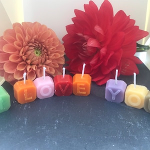Op de afbeelding: Een set kleurrijke kaarsen die "I LOVE YOU" spellen met een rode en oranje bloem op de achtergrond.