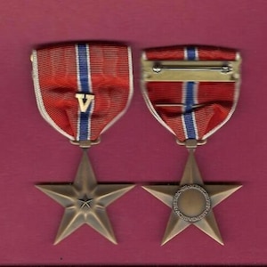 Pode incluir: Uma medalha de estrela de bronze com uma fita vermelha, branca e azul. A medalha tem um design de estrela de cinco pontas com a inscrição "For Valor" no centro. A fita tem um "V" para valor.