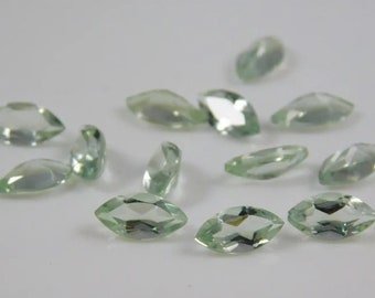 May include: A collection of marquise-cut gemstones in a pale green hue. The gemstones are scattered across a white surface, with varying orientations and angles. The facets of the stones catch the light, creating a sparkling effect. These gems are suitable for jewelry making.