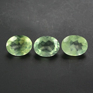 Puede incluir: Tres piedras preciosas ovaladas, de color verde pálido, se muestran sobre un fondo oscuro. Las gemas tienen un aspecto facetado, captando la luz y mostrando su estructura interna. Las piedras están dispuestas en línea horizontal.