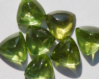 Puede incluir: Una colección de piedras preciosas triangulares translúcidas de color verde oliva. Las piedras facetadas están dispuestas sobre un fondo claro, con la luz reflejándose en sus superficies. Las gemas tienen un aspecto pulido.