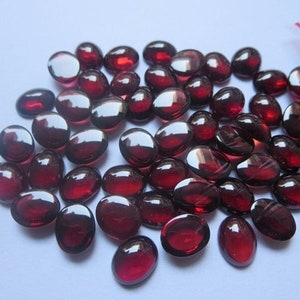 May include: A collection of polished, oval-shaped, deep red gemstones. The stones have a smooth, glossy surface and vary slightly in size. They are scattered on a light background, with a few visible in a small plastic bag.