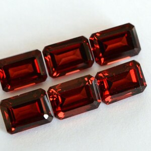 Könnte beinhalten: Sechs rechteckig geschliffene, tiefrote Edelsteine liegen auf einer weißen Oberfläche. Jeder Stein hat eine facettierte Oberfläche, die das Licht reflektiert. Die Steine sind von einem satten, dunklen Rot und haben ein poliertes Aussehen. Wahrscheinlich Granate.