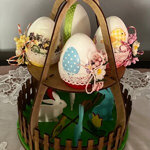 Puede incluir: Un soporte de huevos de Pascua de madera con un diseño de valla y una base verde. Presenta cuatro huevos decorados con cintas y flores. El soporte tiene dos niveles y está decorado con conejos y pollitos.