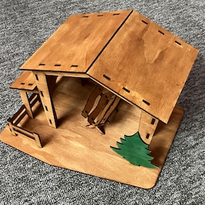 Può includere: Modello in legno di un fienile con un albero verde, una recinzione e una panca all'interno. Il fienile ha una finitura colorata di marrone e un tetto a due falde.