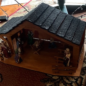 May include: A wooden nativity scene with a black shingled roof. The scene includes figures of Mary, Joseph, baby Jesus, an angel, a shepherd, and a donkey. The figures are made of painted ceramic.
