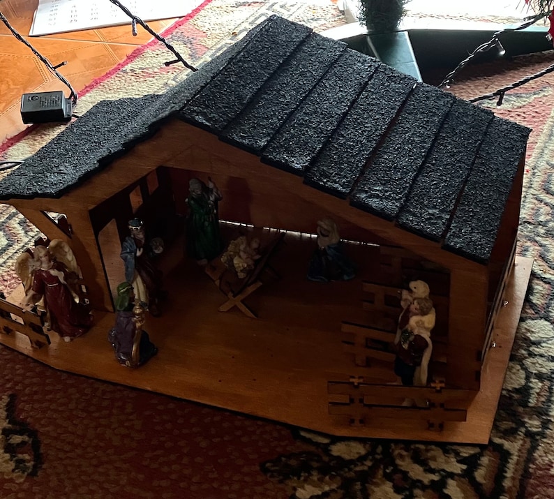 May include: A wooden nativity scene with a black shingled roof. The scene includes figures of Mary, Joseph, baby Jesus, an angel, a shepherd, and a donkey. The figures are made of painted ceramic.