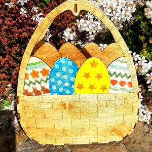 Puede incluir: Cesta de huevos de Pascua de madera con un diseño recortado. La cesta está pintada de marrón y tiene cuatro huevos coloridos en su interior. Los huevos están pintados con diferentes patrones, incluyendo lunares, estrellas y corazones.