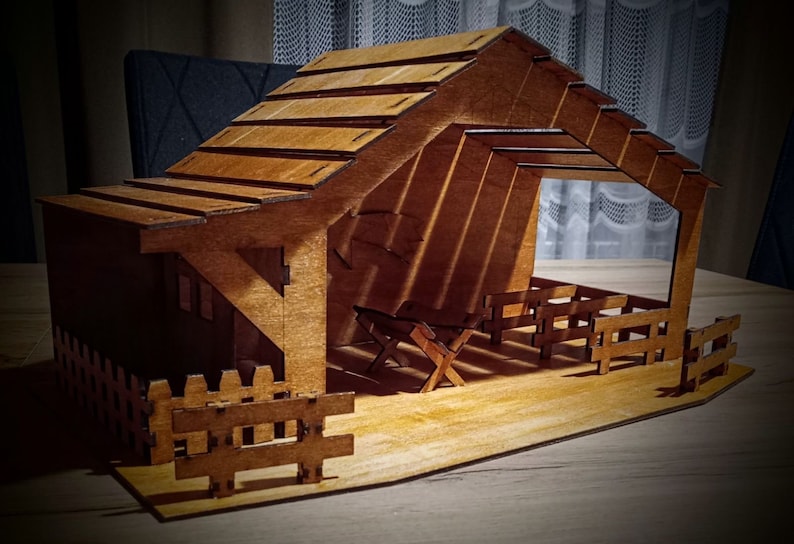 May include: A wooden nativity scene with a brown wooden stable, a fence, and two wooden benches inside. The stable has a roof with a slanted design.