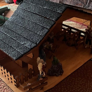 May include: A wooden nativity scene with a black shingled roof, a brown wooden fence, and figurines of Mary, Joseph, and the baby Jesus. The scene is set in a stable with a manger and hay.