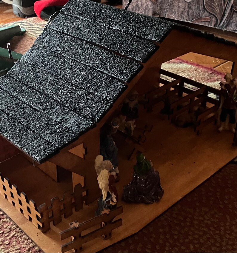 May include: A wooden nativity scene with a black shingled roof, a brown wooden fence, and figurines of Mary, Joseph, and the baby Jesus. The scene is set in a stable with a manger and hay.