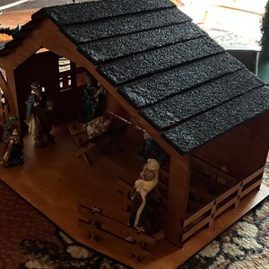 May include: A wooden nativity scene with a black shingled roof. The scene includes figures of Mary, Joseph, baby Jesus, and a shepherd. The figures are made of painted plastic and are standing on a wooden base.
