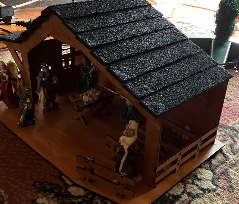 May include: A wooden nativity scene with a black shingled roof. The scene includes figures of Mary, Joseph, baby Jesus, and a shepherd. The figures are made of painted plastic and are standing on a wooden base.