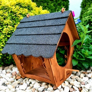 Puede incluir: Un comedero para pájaros de madera con un techo de tejas negras. El comedero tiene una pequeña plataforma y un diseño decorativo de corte.