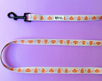 May include: A gray and white dog leash with a gingerbread house pattern. The leash has a black metal clip and a white label that says "House of Hugo".