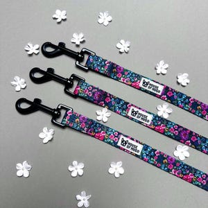 Puede incluir: Tres correas para perros negras con tela de estampado floral y clips de metal negro. Las correas tienen el texto "House of Hugo" impreso en ellas.