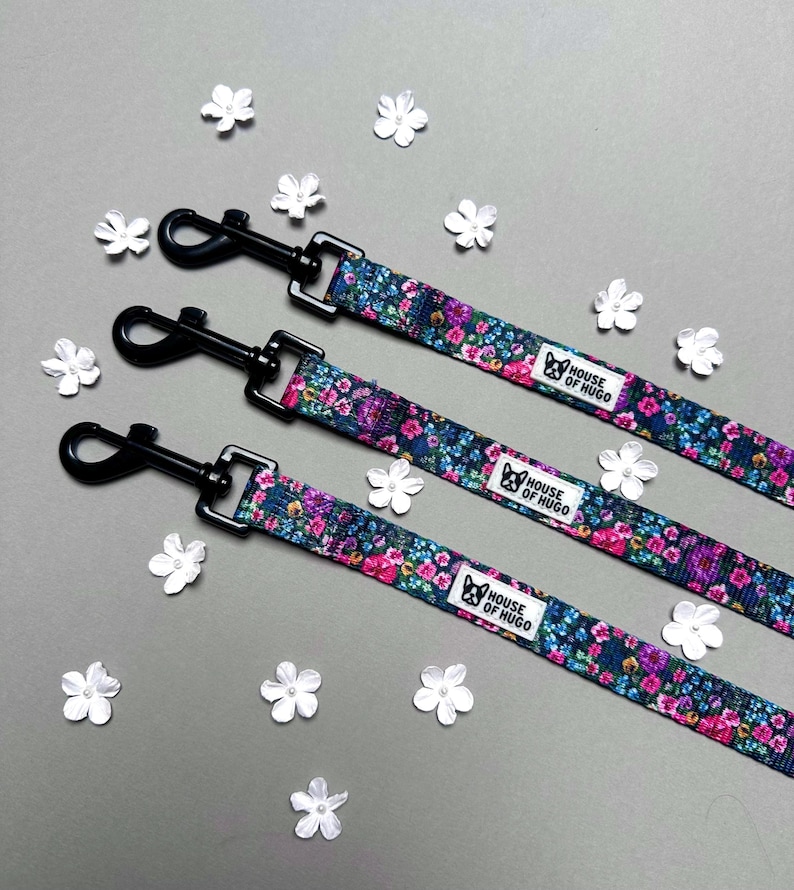 Puede incluir: Tres correas para perros negras con tela de estampado floral y clips de metal negro. Las correas tienen el texto "House of Hugo" impreso en ellas.