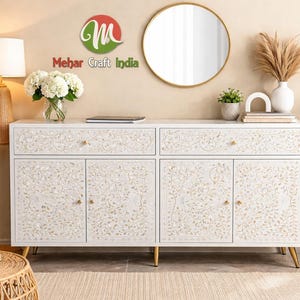 Puede incluir: Aparador de madera blanca con diseño de incrustaciones florales, herrajes y patas doradas. El mueble tiene dos cajones y cuatro puertas. La imagen también muestra un espejo redondo, plantas y objetos decorativos. El logo "Mehar Craft India" es visible.