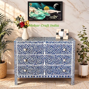 Op de afbeelding: Blauwe dressoir met een wit bloemmotief. De dressoir heeft vier lades en witte knoppen. De tekst "Mehar Craft India" is zichtbaar. Een ingelijste foto en een vaas met bloemen staan erop.
