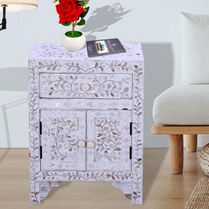 May include: White nightstand with a floral design made of mother of pearl inlay. The nightstand has a drawer and two doors. A red rose is on top of the nightstand.