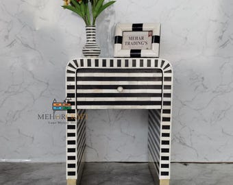 May include: A black and white striped nightstand with a single drawer and gold-colored feet. The nightstand has a vase of flowers and a framed picture on top. The picture frame and vase have matching black and white stripes. The text "MEHAR TRADING'S" is visible on the picture frame.