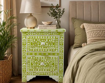 May include: A lime green bedside table featuring two drawers with intricate white floral inlay patterns. The rectangular piece has a flat top surface and stands on a base with a scalloped cutout design. The surface is decorated with white leaf motifs.