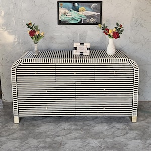 Può includere: Una cassettiera a righe bianche e nere con sei cassetti. La cassettiera ha un design curvo e piedini color oro. Due vasi con fiori sono posti sopra, insieme a un'insegna incorniciata con la scritta "MEHAR TRADING'S". Un'immagine incorniciata è appesa al muro sopra.