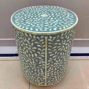 May include: A cylindrical side table featuring a grey base with an intricate inlaid pattern of white leaf motifs. The top surface displays a symmetrical floral design, while the sides are decorated with vertical vine patterns in white inlay.