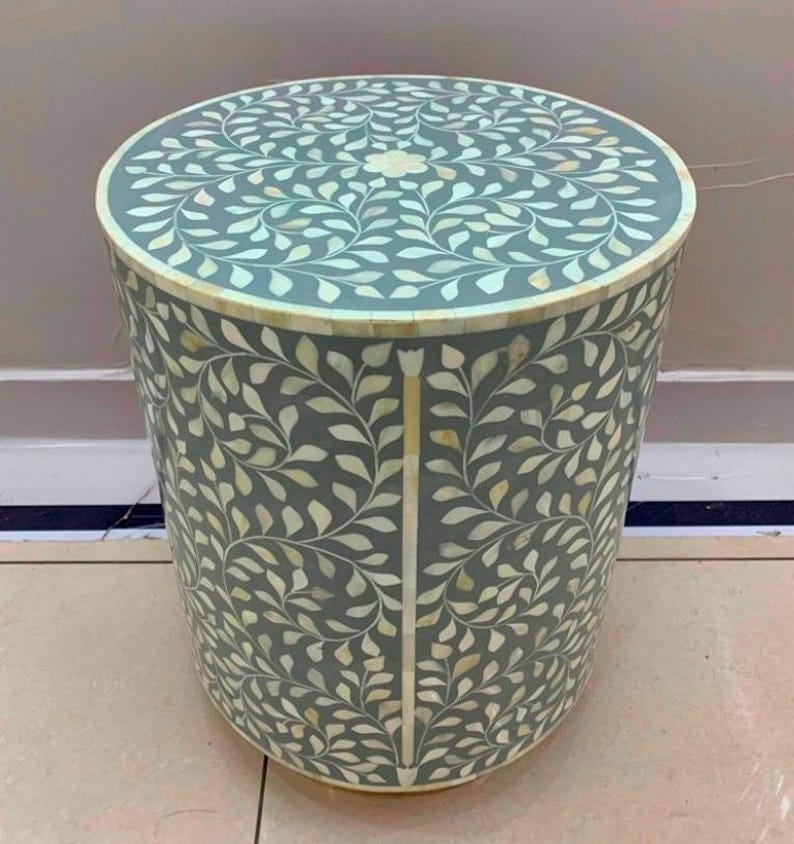May include: A cylindrical side table featuring a grey base with an intricate inlaid pattern of white leaf motifs. The top surface displays a symmetrical floral design, while the sides are decorated with vertical vine patterns in white inlay.
