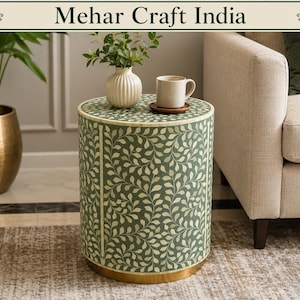 May include: A cylindrical side table featuring a sage green surface with an intricate white leaf vine pattern. The table has a gold-toned metal base and a smooth top surface holding a white ribbed vase with greenery and a beige ceramic mug.
