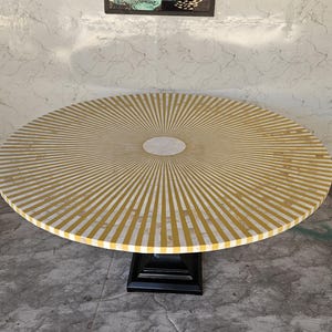 May include: A round table with a sunburst design. The tabletop features alternating yellow and white stripes radiating from a central white circle. The table is supported by a black, square pedestal base.