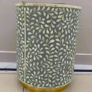 May include: A cylindrical side table featuring a grey surface inlaid with a cream-colored leaf and vine pattern made of bone. The table has a circular gold-toned metal base and a flat top edge, showcasing a repeating botanical motif throughout.