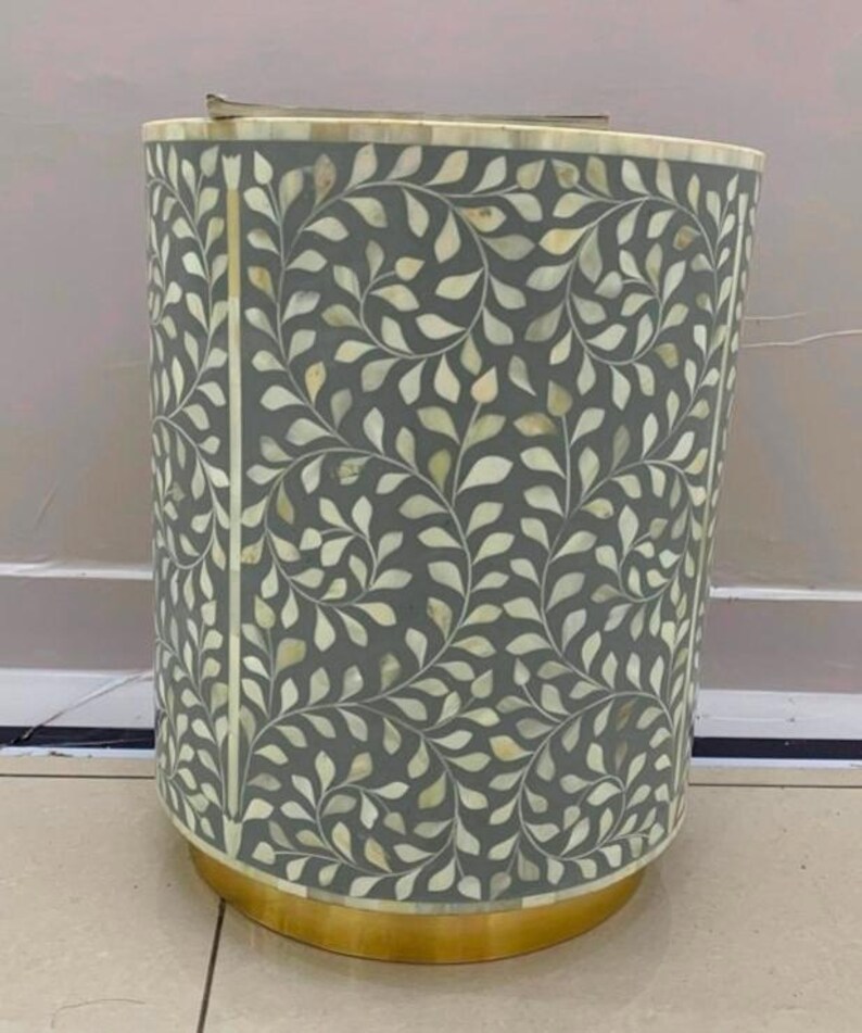 May include: A cylindrical side table featuring a grey surface inlaid with a cream-colored leaf and vine pattern made of bone. The table has a circular gold-toned metal base and a flat top edge, showcasing a repeating botanical motif throughout.