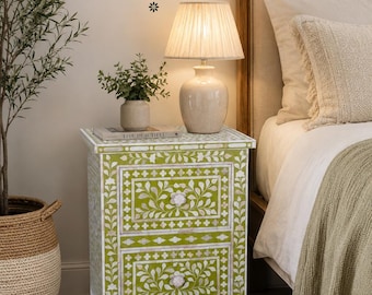 May include: A lime green bedside table with two drawers featuring white floral mother-of-pearl inlay patterns. The text mehar craft india, CRAFTED WITH HERITAGE, MADE FOR YOUR HOME. is visible above the table, which holds a beige lamp and plant.