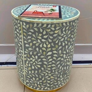 May include: A cylindrical side table with a grey surface featuring an intricate white leaf pattern inlay. A rectangular brochure with the text MEHAR TRADING'S is placed on top. The base of the table is finished with a metallic gold-toned trim.