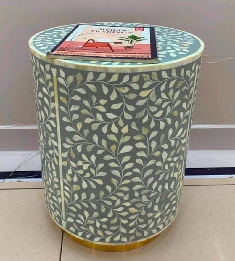 May include: A cylindrical side table with a grey surface featuring an intricate white leaf pattern inlay. A rectangular brochure with the text MEHAR TRADING'S is placed on top. The base of the table is finished with a metallic gold-toned trim.