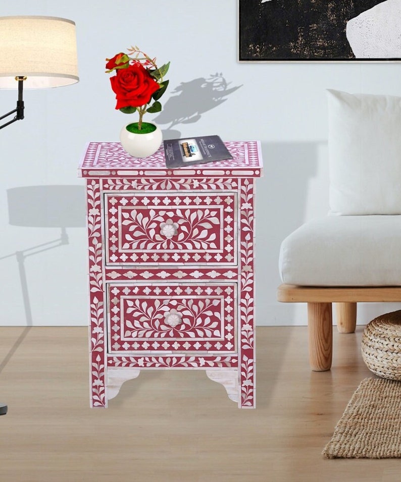 May include: A two-drawer nightstand with a pink and white bone inlay design. The drawers have floral patterns and white knobs. The nightstand is sitting on a wooden floor.