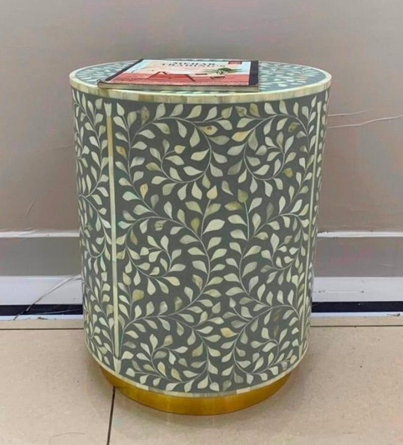 May include: A cylindrical side table featuring a grey surface with an intricate white leaf pattern inlay. The table has a solid gold-colored circular base and a flat top surface currently holding a rectangular book with a red and white cover design.