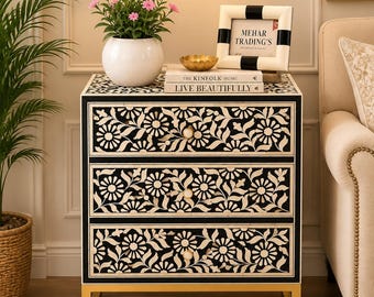 May include: A three-drawer chest featuring a black and white floral inlay pattern on the drawer fronts. The chest has a rectangular frame with a gold-toned metal base. The top surface holds a white vase with pink flowers and a picture frame.