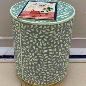 May include: A cylindrical side table with a grey base and an intricate white leaf pattern inlay. A rectangular catalog titled MEHAR TRADING'S is placed on the flat top surface. The base of the table features a solid gold-colored metal trim.
