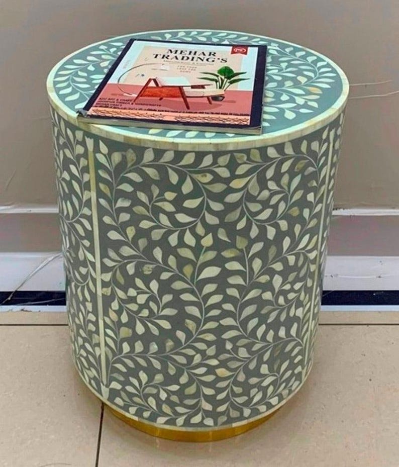 May include: A cylindrical side table with a grey base and an intricate white leaf pattern inlay. A rectangular catalog titled MEHAR TRADING'S is placed on the flat top surface. The base of the table features a solid gold-colored metal trim.