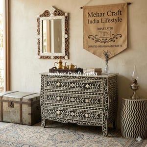 May include: A black and white floral inlay chest of drawers with three drawers, a matching mirror, and a decorative side table. The room is decorated with a vintage trunk, rug, and a banner that reads "Mehar Craft India Lifestyle."