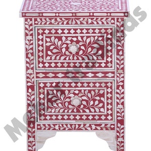 May include: A red wooden nightstand with two drawers. The drawers are decorated with white floral patterns and intricate bone inlay. The nightstand has a traditional design and is perfect for adding a touch of elegance to any bedroom.