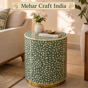 May include: A cylindrical side table featuring a green surface with an intricate white leaf pattern, a gold-toned metal base, and the text Mehar Craft India at the top. The table holds a white vase with flowers and a book titled Kinfolk.