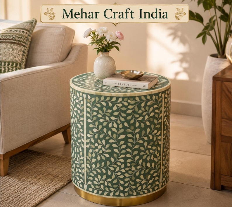 May include: A cylindrical side table featuring a green surface with an intricate white leaf pattern, a gold-toned metal base, and the text Mehar Craft India at the top. The table holds a white vase with flowers and a book titled Kinfolk.