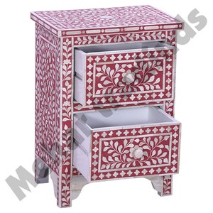 May include: A red wooden nightstand with two drawers. The drawers are decorated with white floral patterns and have bone inlay details. The nightstand has a floral design on the front and sides.